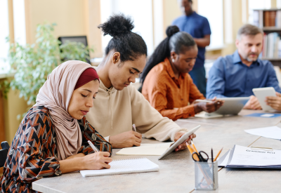 Gruppo di persone di etnia diversa che svolgono una lezione di lingua per immigrati seduti a un tavolo che svolgono attività con l'uso di tablet digitali